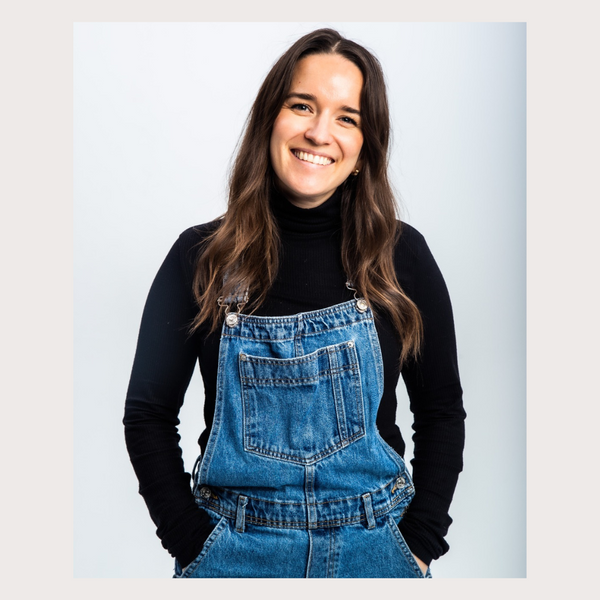 fiona fernie woman with long dark brown hair smiling with her mouth open looking straight at the camera wearing black long sleeve roll neck and blue jean overalls with hands in her pockets