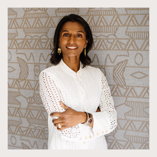Image of the top half of a  dark skinned woman standing, smiling against a grey decorated wall, wearing a white long sleeved top, gold jewellery with her arms crossed.