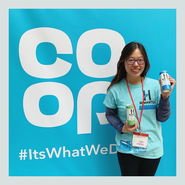 Image of Tina Chen Woman with Asian features long dark hair, glasses, wearing a turquoise tee shirt and a lanyard holding up two can of HumaniTea with black trousers and standing in front of a turquoise sign with white lettering that reads coop