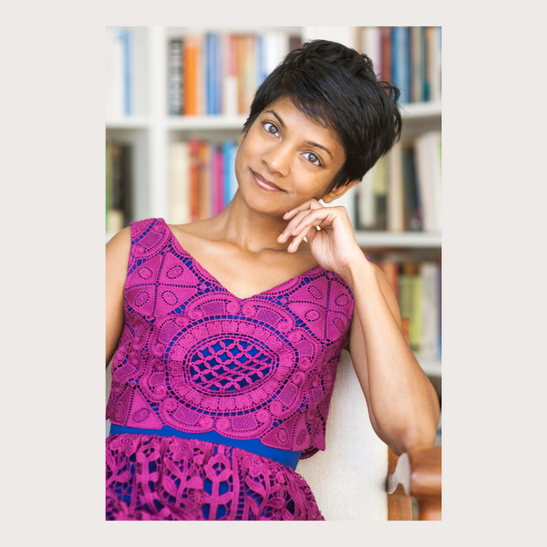 image of vena ramphal south asian woman with short dark hair, seated with head tilted to one side, wearing sleeveless magenta and blue v neck dress, smiling with lips closed with a background of bookshelves.