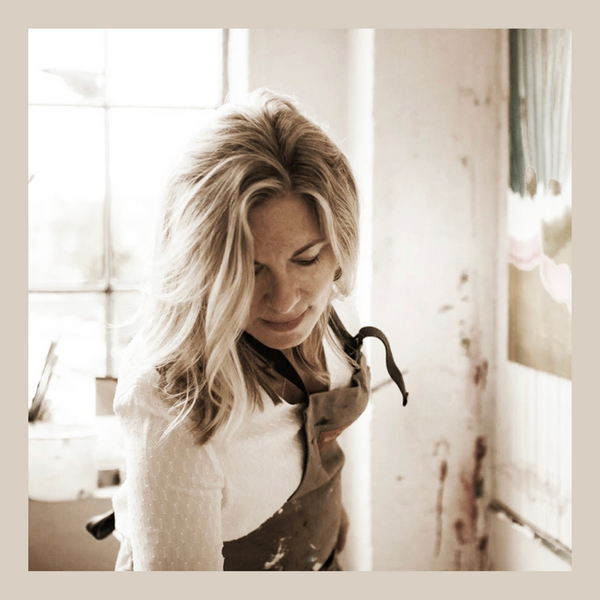 image of Kate Lowe, artist, looking down, with a window behind her, wearing a white top and apron, with blonde shoulder length hair and a painting to her left on the wall