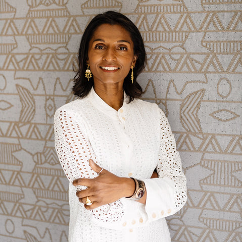 Image of the top half of a  dark skinned woman standing, smiling against a grey decorated wall, wearing a white long sleeved top, gold jewellery with her arms crossed.