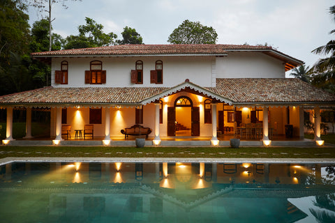 image of a large, white two storied house with red roof tiles. The ground floor is lit up with interior and exterior lights and a large turquoise pool is in front of the house. The house is set against greenery and a pale blue sky with some clouds.
